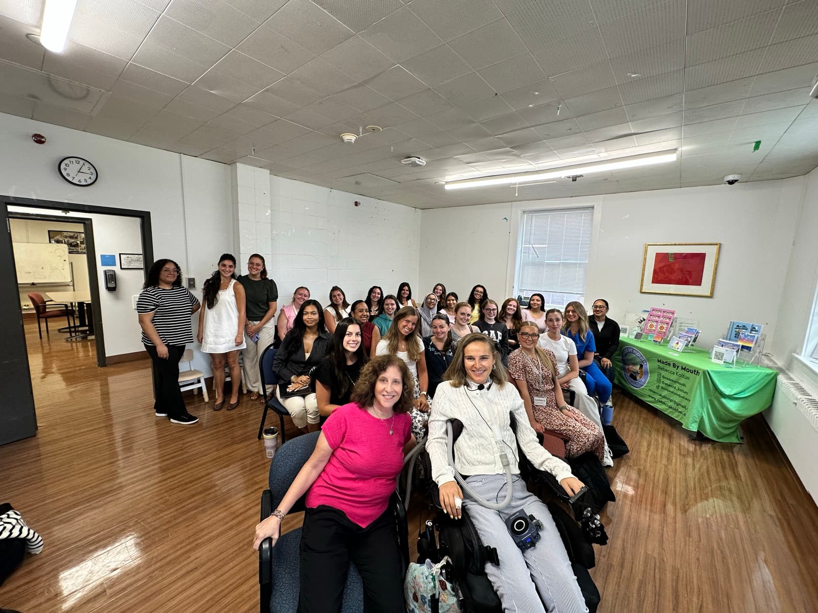 Rebecca in her wheelchair at the front of a classroom, surrounded by a smiling group of speech-language pathology students at Long Island University.