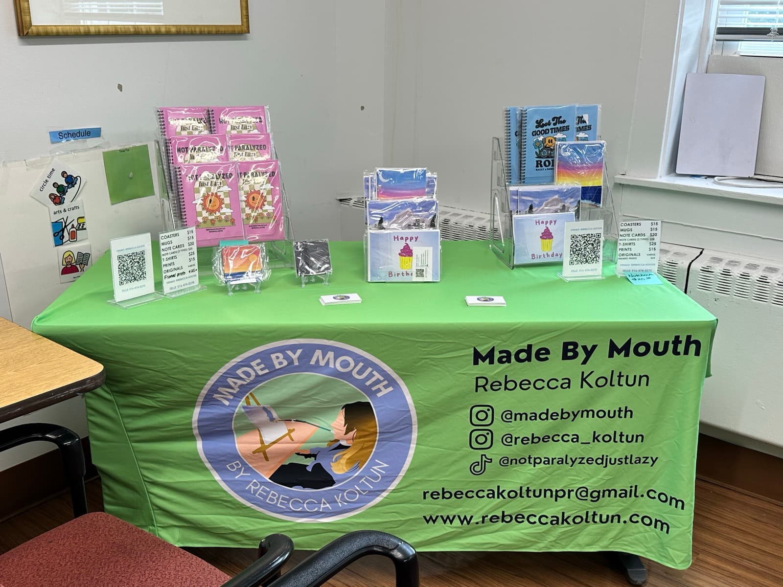 The Made by Mouth table at LIU: notebooks, note cards, coasters and prints of Rebecca's mouth paintings on a green tablecloth.