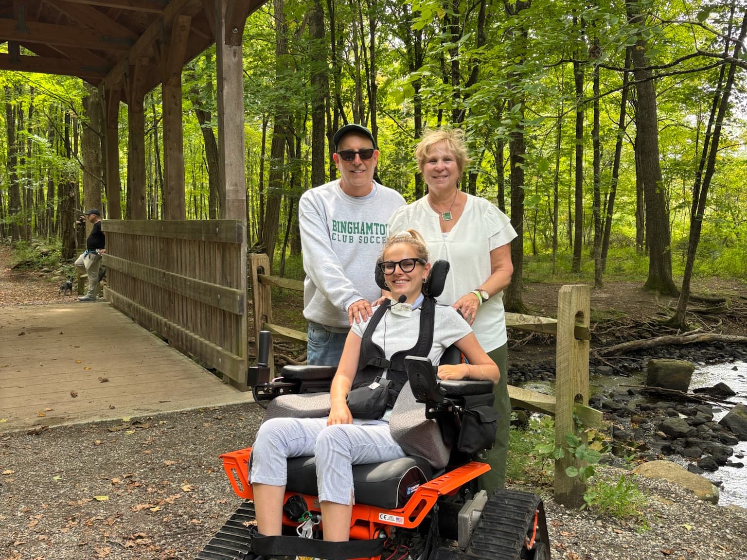 Rebecca in an all-terrain track wheelchair with her parents by a covered bridge on a wooded trail in Upstate New York.